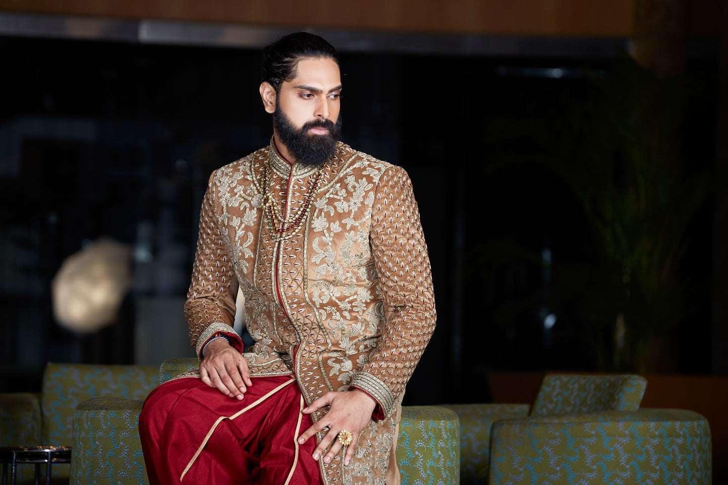 A man wearing a traditional Indian sherwani and dhoti outfit, featuring a beige and gold patterned sherwani with red dhoti pants, seated in a dimly lit indoor setting.