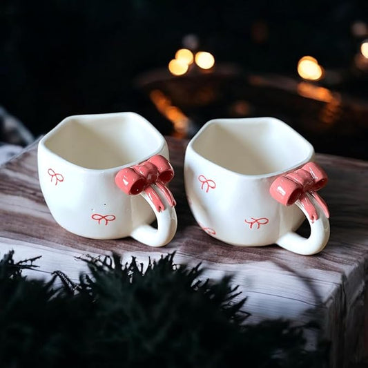 Two white mugs with red bow handles on a wooden surface with a blurred background.