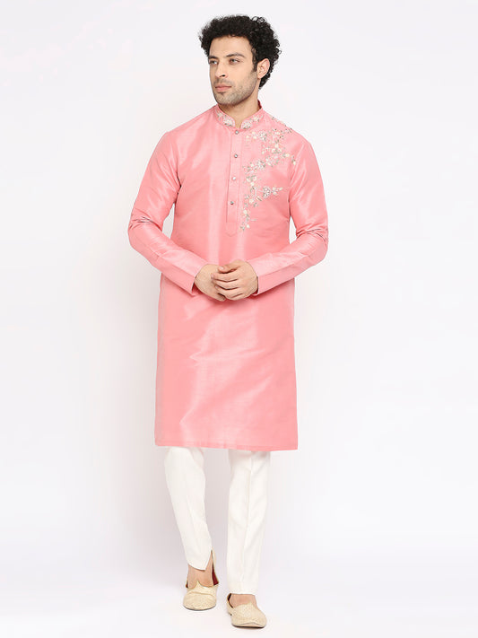 A man modeling a light pink long kurta with silver floral embroidery on the chest, paired with white trousers and beige shoes, against a plain white background.
