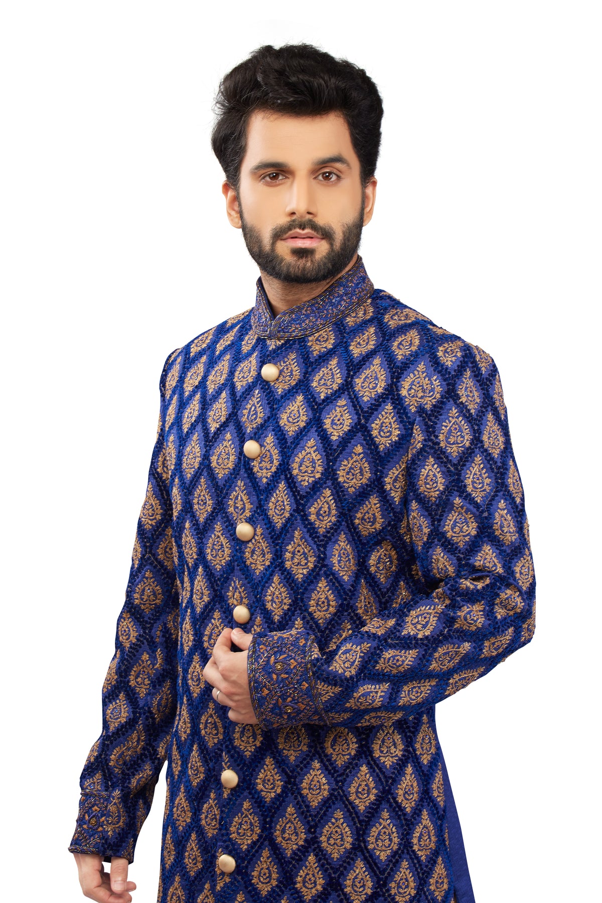 A man wearing a royal blue sherwani jacket with an intricate gold and brown geometric pattern, featuring a mandarin collar and decorative buttons, set against a plain white background.