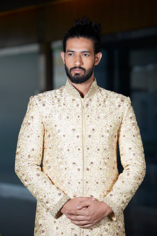 A man wearing a cream-colored sherwani jacket with intricate gold and brown floral embroidery, standing against a dark, blurred background.