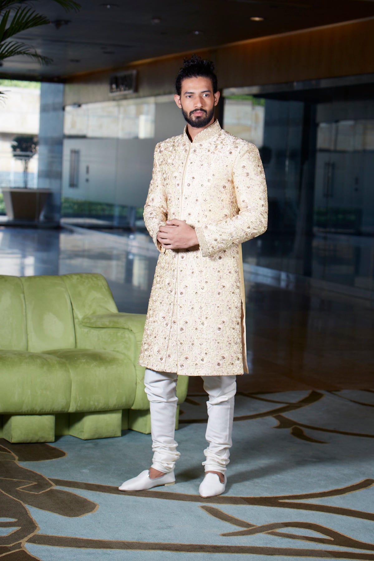A man wearing a cream-colored, long-sleeved sherwani with gold floral embroidery, paired with white trousers and white shoes, standing in a modern lobby with a green sofa in the background.