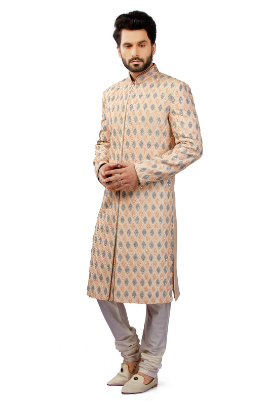 A man modeling a beige and gray patterned sherwani long coat with light gray trousers and beige shoes, standing against a plain white background.