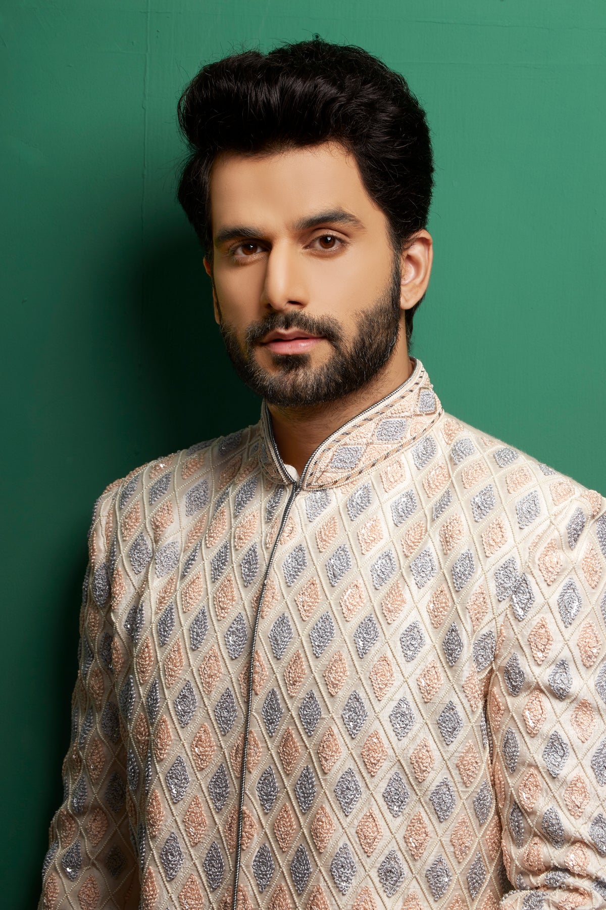A man wearing a beige sherwani jacket with a blue and pink geometric pattern, posed against a solid green background.
