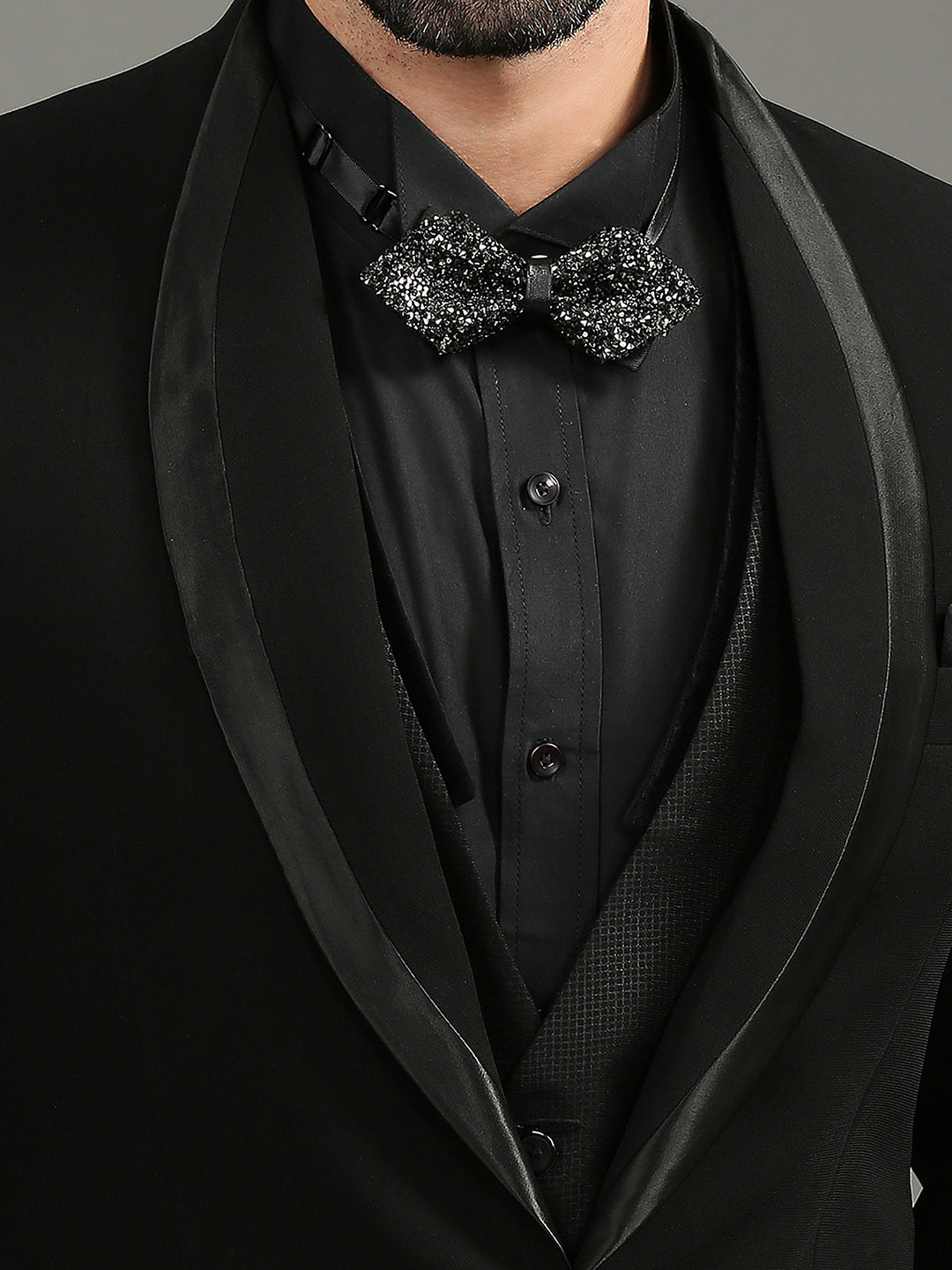 A close-up of a man wearing a black tuxedo jacket with satin shawl lapels, a black dress shirt, a black vest, and a black sequined bow tie.