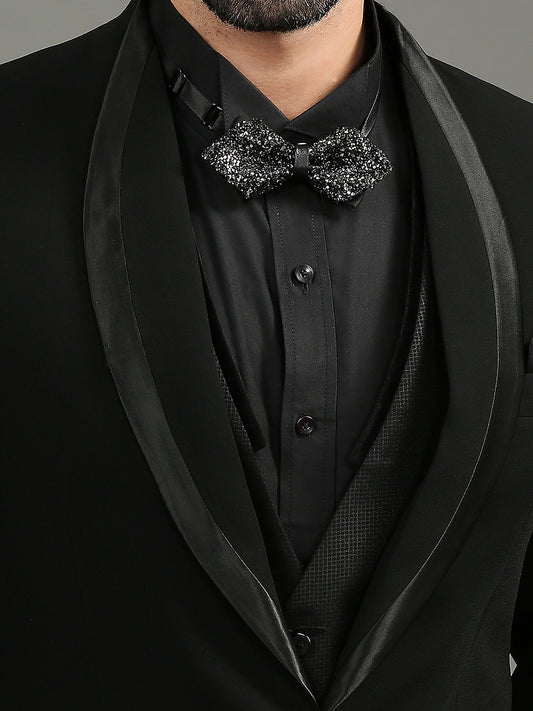 A close-up of a man wearing a black tuxedo jacket with satin shawl lapels, a black dress shirt, a black vest, and a black sequined bow tie.