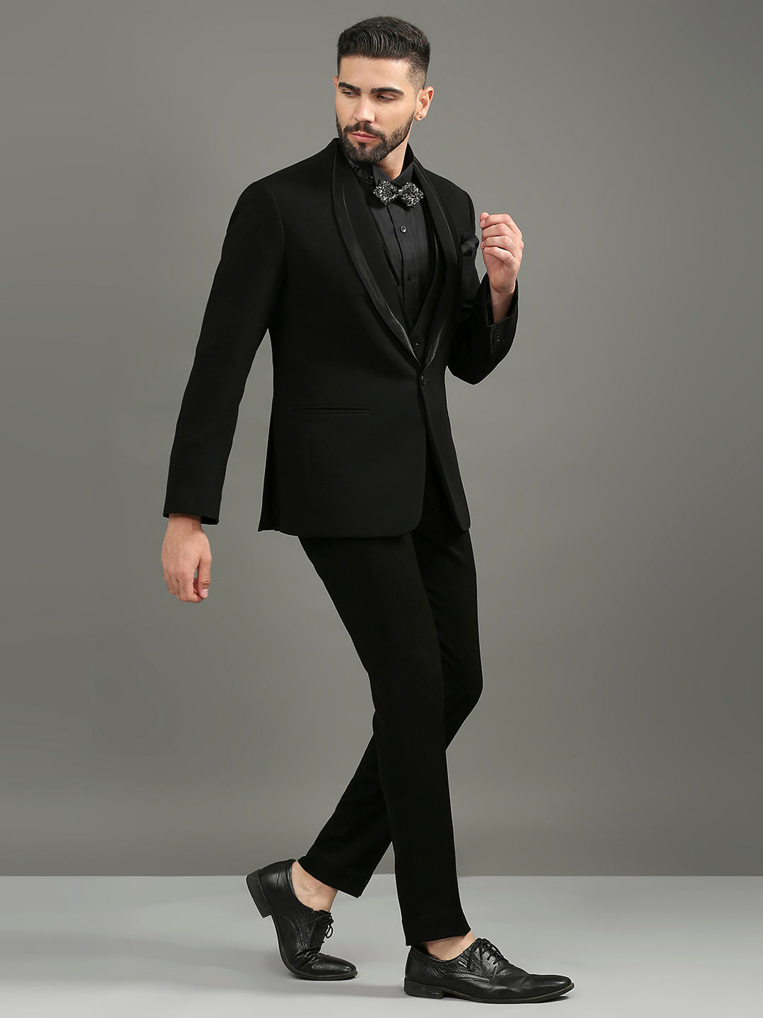 A man modeling a black formal tuxedo with satin peak lapels, a black shirt, a black bow tie, and black dress shoes against a solid gray studio background.
