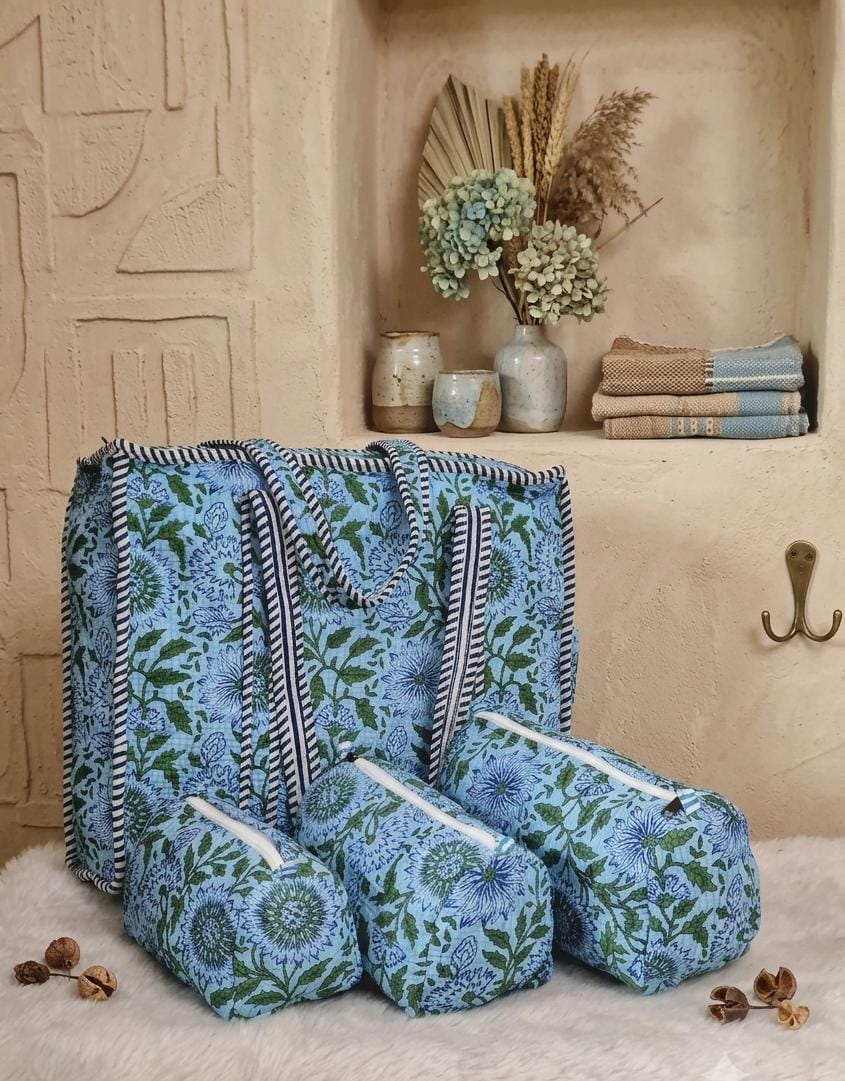 A matching fabric storage set including a large tote bag and three zippered pouches, all featuring a blue and green floral pattern with striped handles and trim, styled on a white faux fur rug in front of a beige plaster wall.