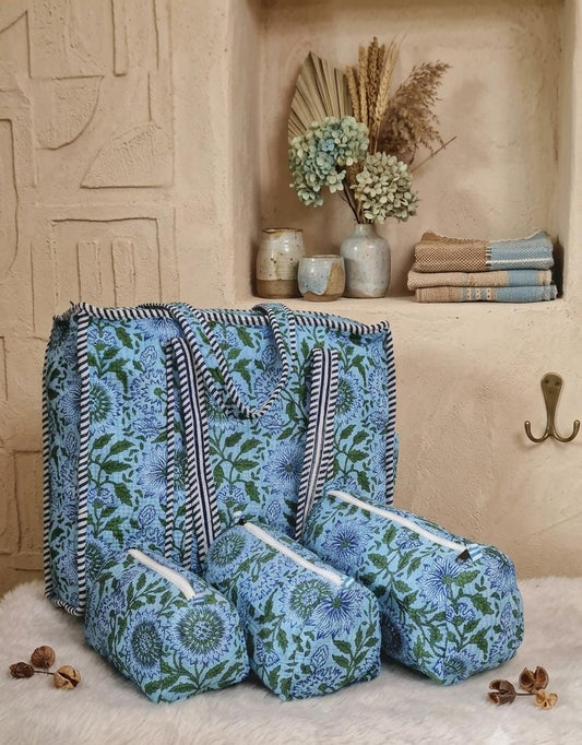 A matching fabric storage set including a large tote bag and three zippered pouches, all featuring a blue and green floral pattern with striped handles and trim, styled on a white faux fur rug in front of a beige plaster wall.