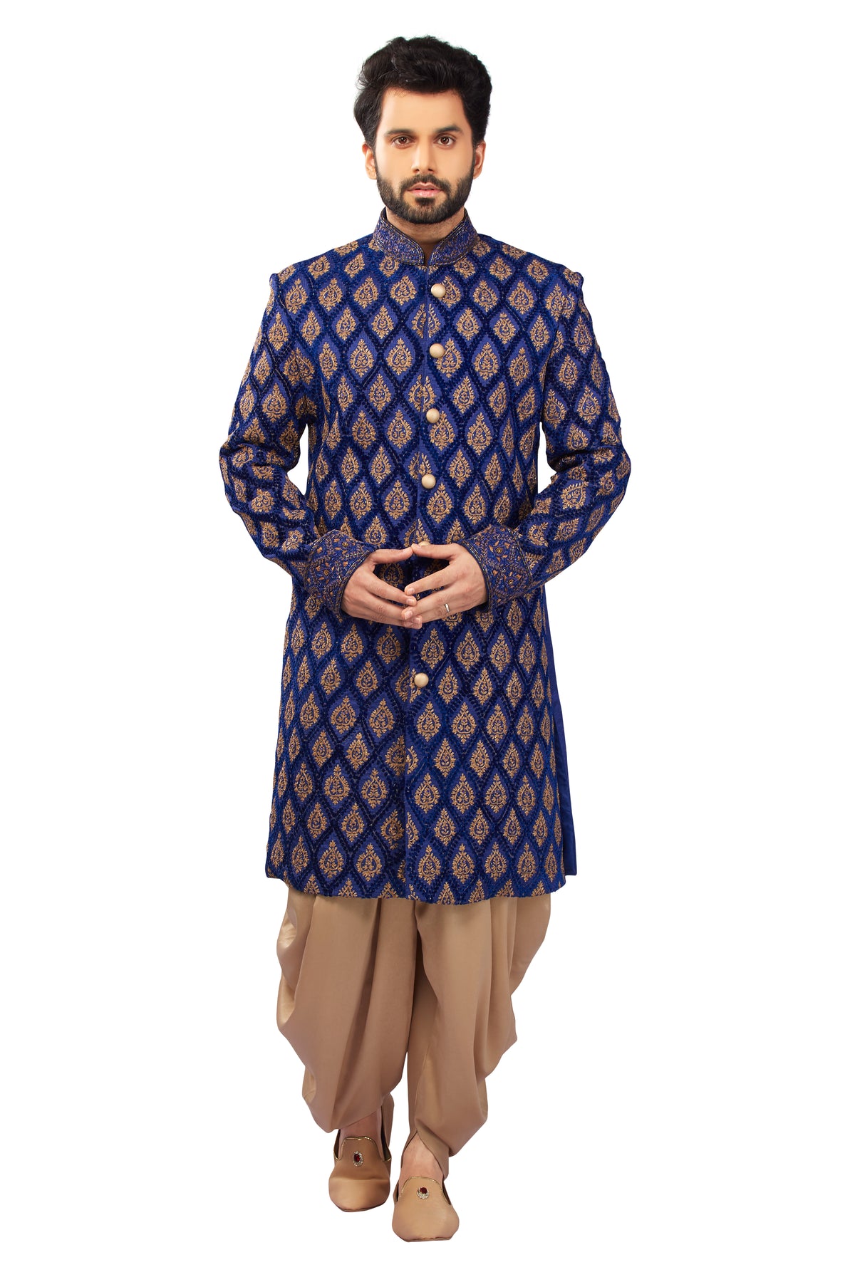 A man modeling a traditional Indian outfit consisting of a royal blue patterned sherwani jacket with gold motifs, paired with beige dhoti pants and brown shoes, against a solid white background.