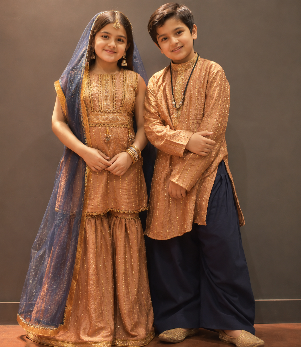 A young boy and girl modeling traditional South Asian clothing. The girl wears a peach-beige embroidered kurta and sharara pants with a blue dupatta, while the boy wears a matching peach-beige embroidered kurta with dark navy pants.