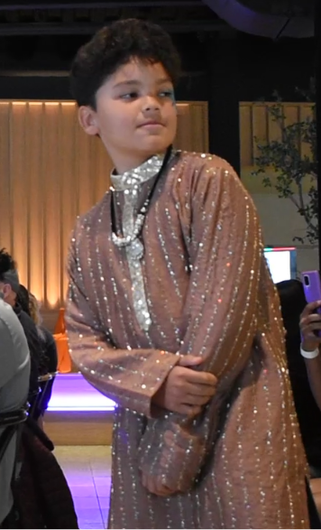 A young boy wearing a sparkling, long-sleeved metallic rose-gold or copper sequined tunic with a silver high collar and layered silver necklaces, standing in an indoor event space.