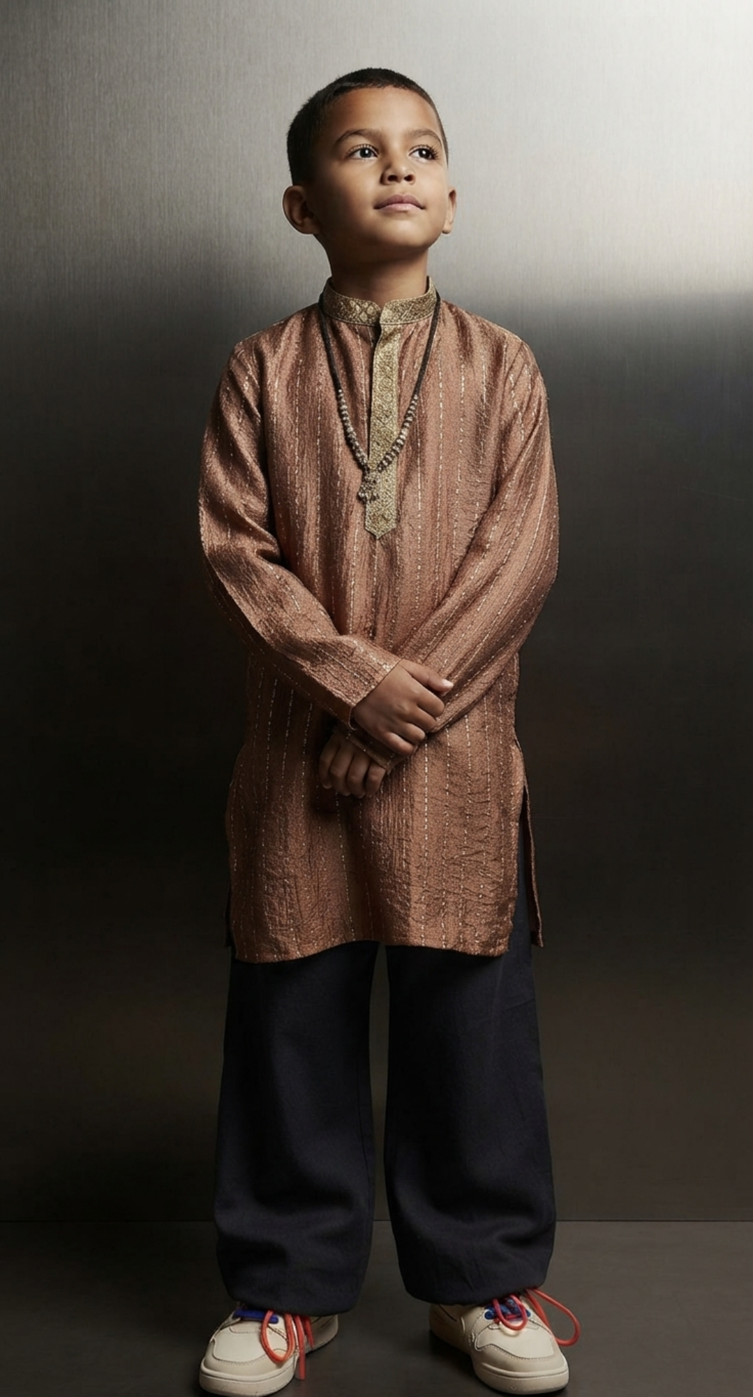 A young boy modeling a traditional Indian outfit consisting of a metallic bronze-brown kurta with gold embroidery, dark navy blue pants, and white sneakers, posed against a dark gray studio backdrop.