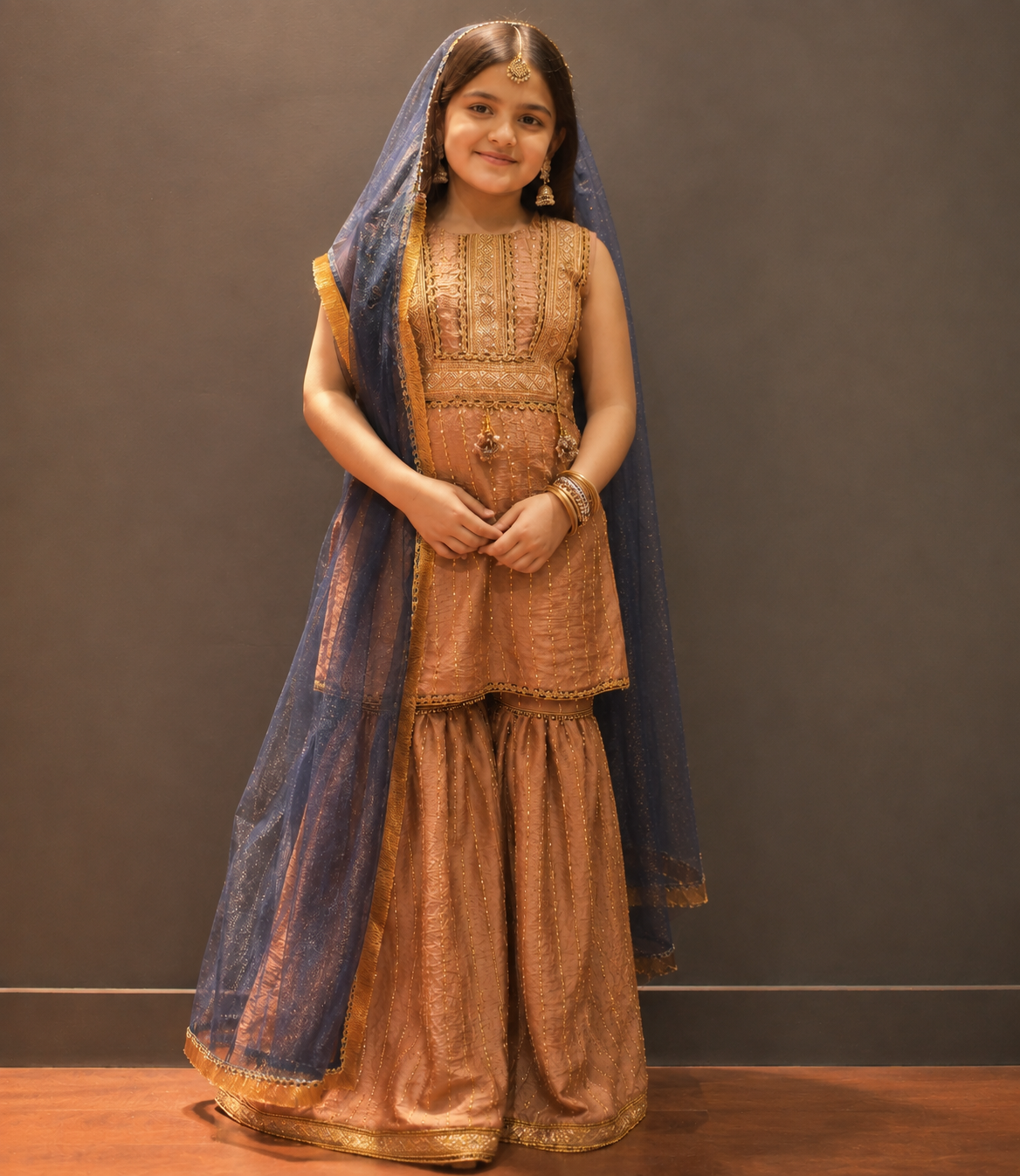 A young girl modeling a traditional Indian outfit consisting of a bronze-gold embroidered kurta, matching flared pants, and a deep blue dupatta with gold borders, set against a dark gray studio background.