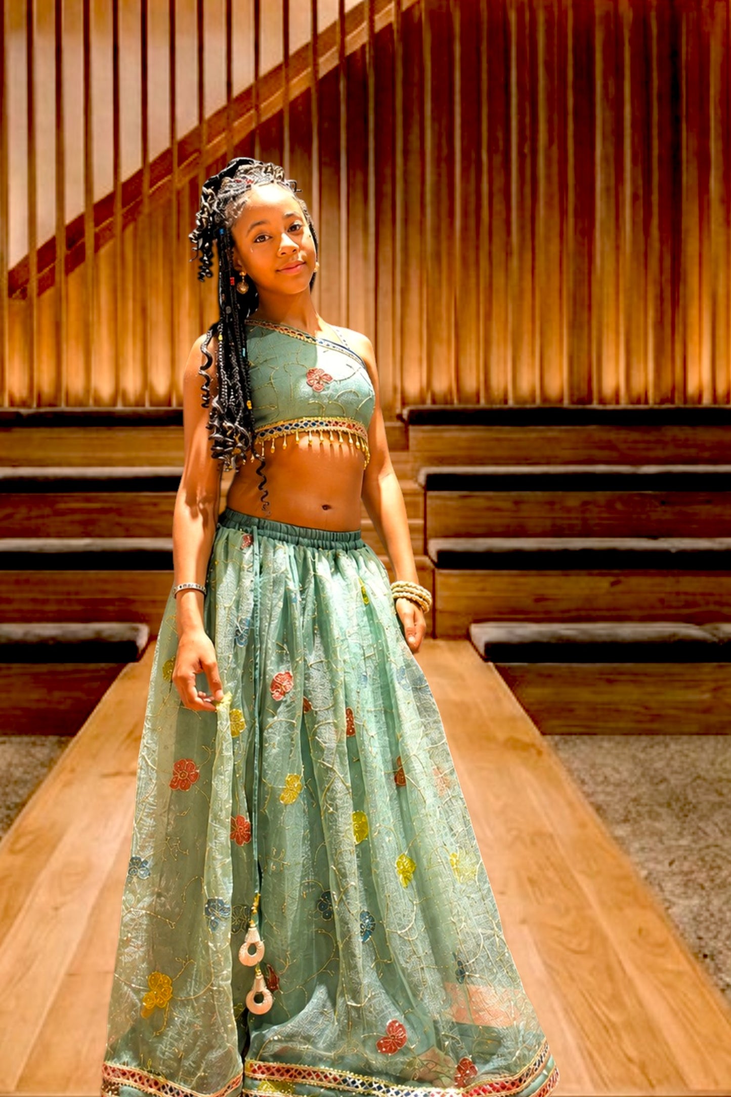 A young girl models a light green two-piece ethnic lehenga choli set with colorful floral embroidery, standing on a wooden runway in front of a wooden slatted wall.