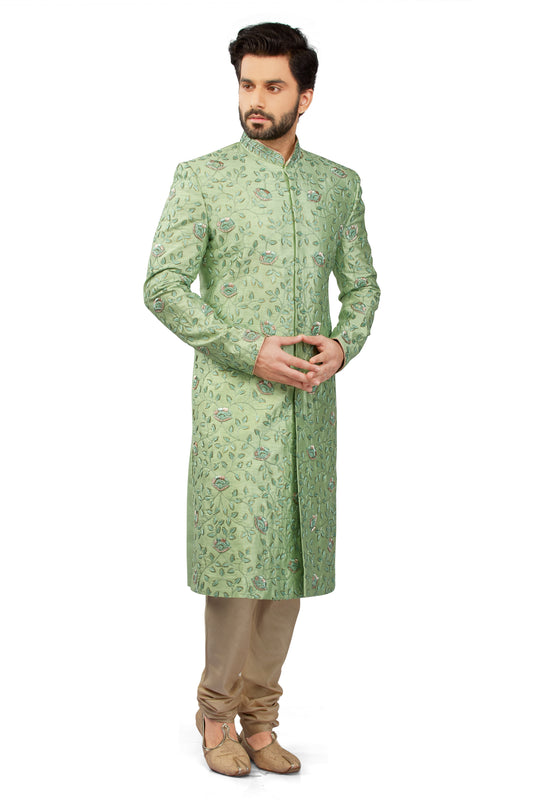 A man modeling a light green long sherwani coat with an intricate paisley pattern, paired with beige trousers and brown shoes, against a plain white background.
