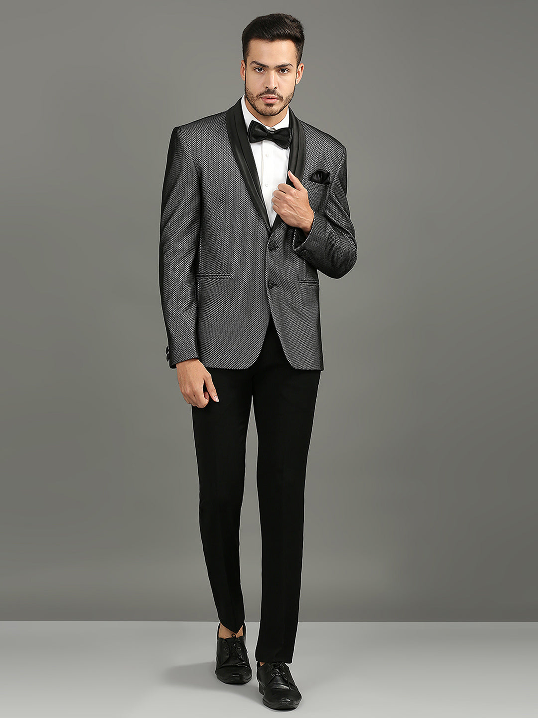 A man modeling a gray textured tuxedo jacket with black satin lapels, paired with black trousers, a white dress shirt, a black bow tie, and a black pocket square, against a solid gray studio background.