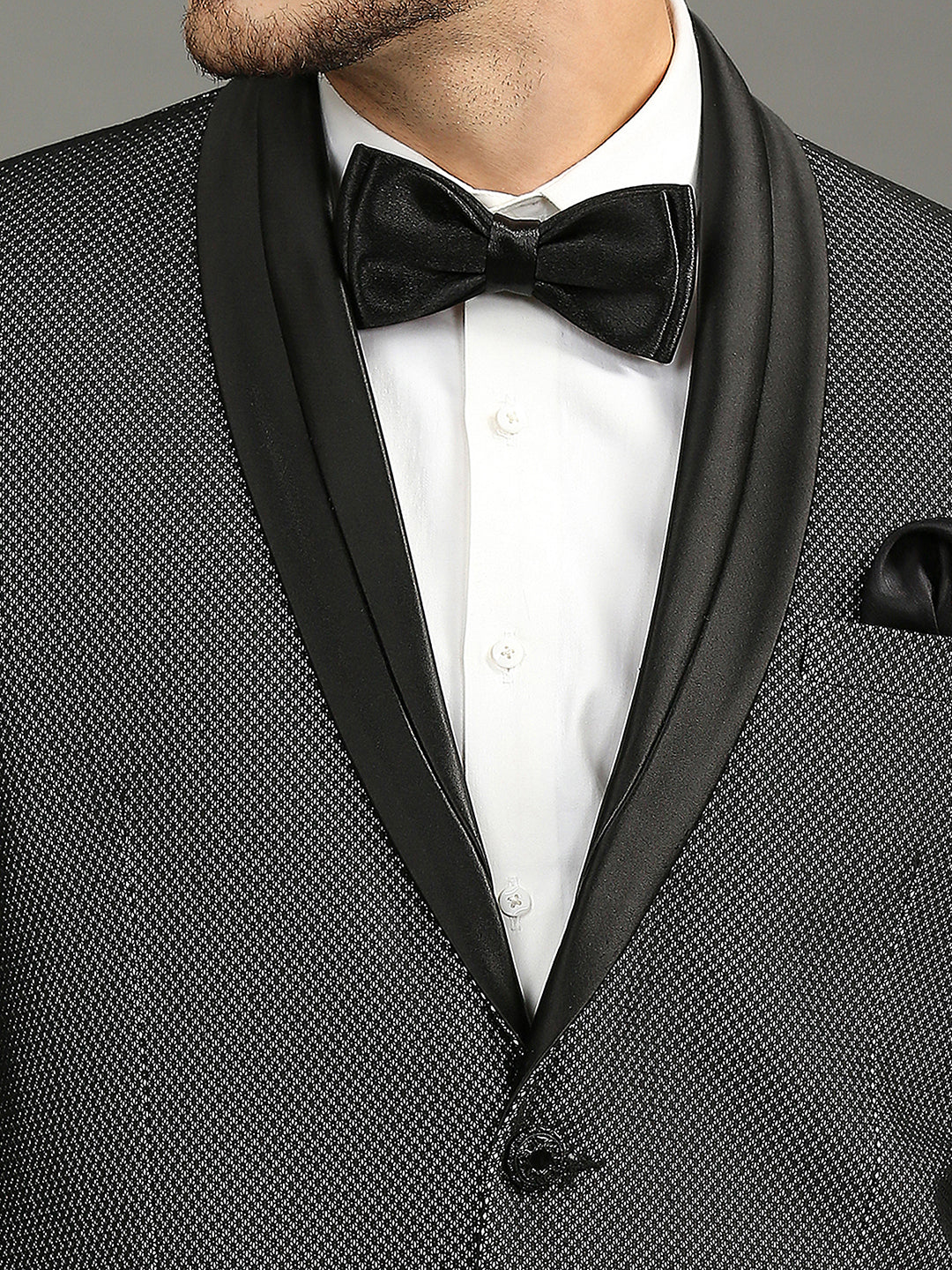 Close-up of a man wearing a black textured tuxedo jacket with satin shawl lapels, a white dress shirt, a black satin bow tie, and a black pocket square.