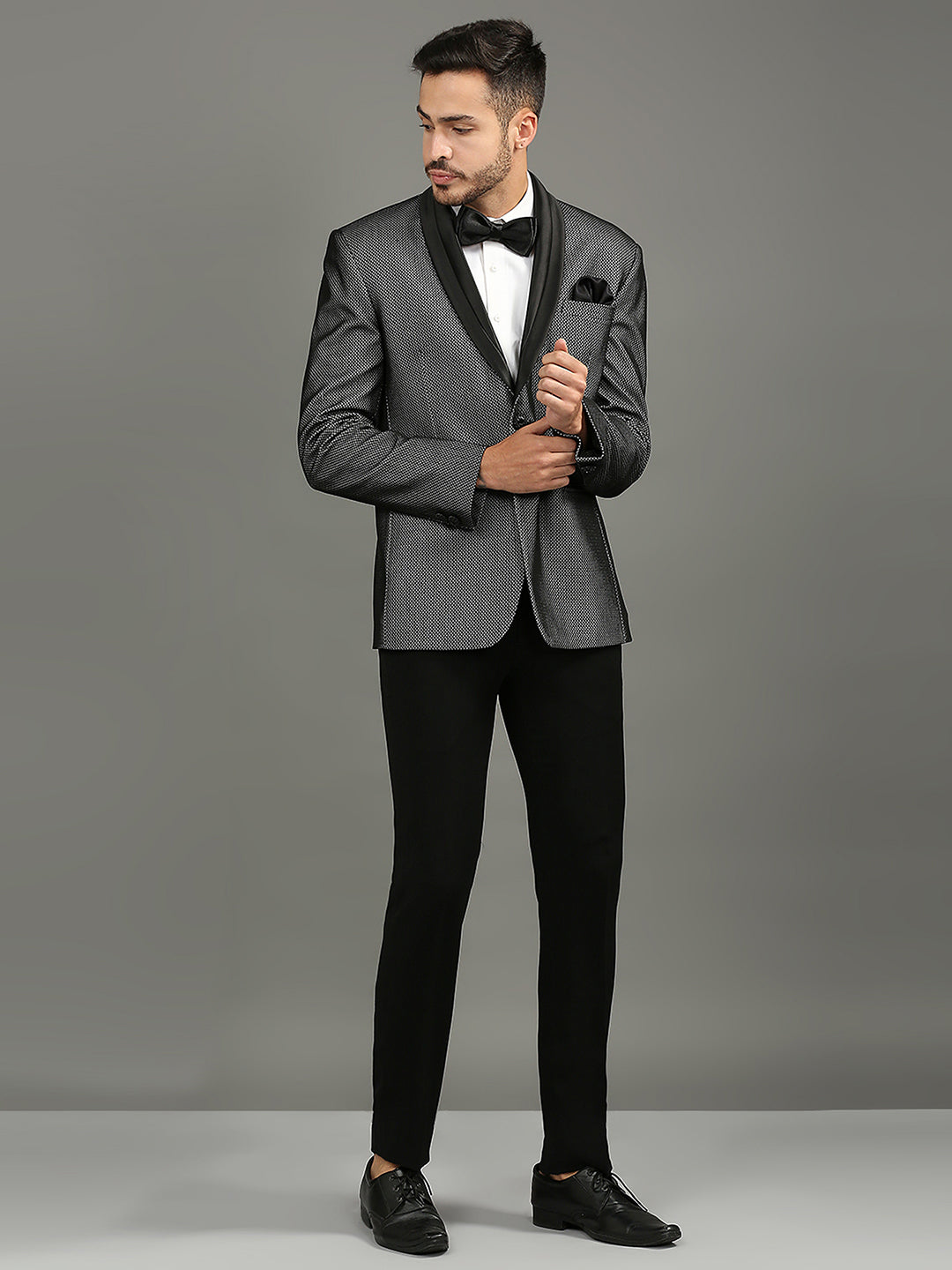 A man modeling a gray tuxedo jacket with black satin lapels, paired with black trousers, a white dress shirt, a black bow tie, and a black pocket square, against a solid gray studio background.