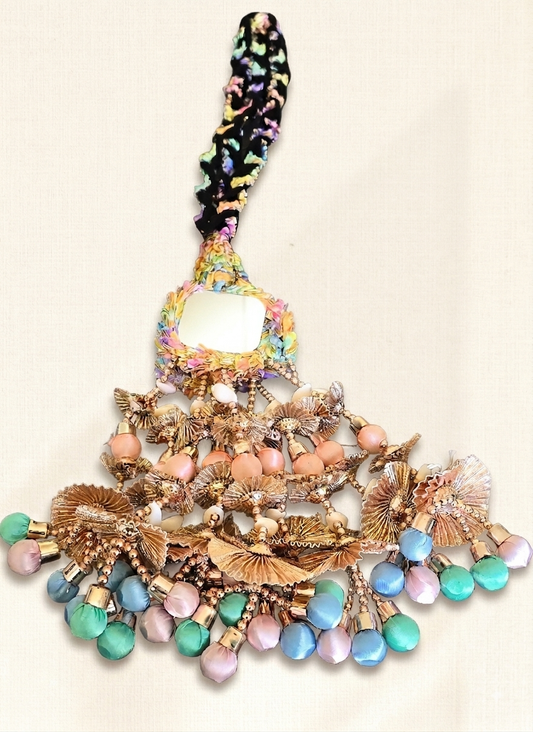 A highly decorative, bohemian-style statement brooch or handbag charm featuring a central mirror, a black patterned top handle, and cascading layers of gold-toned metallic elements, iridescent beads, and pastel-colored glass beads in pink, blue, and green on a light beige fabric background.