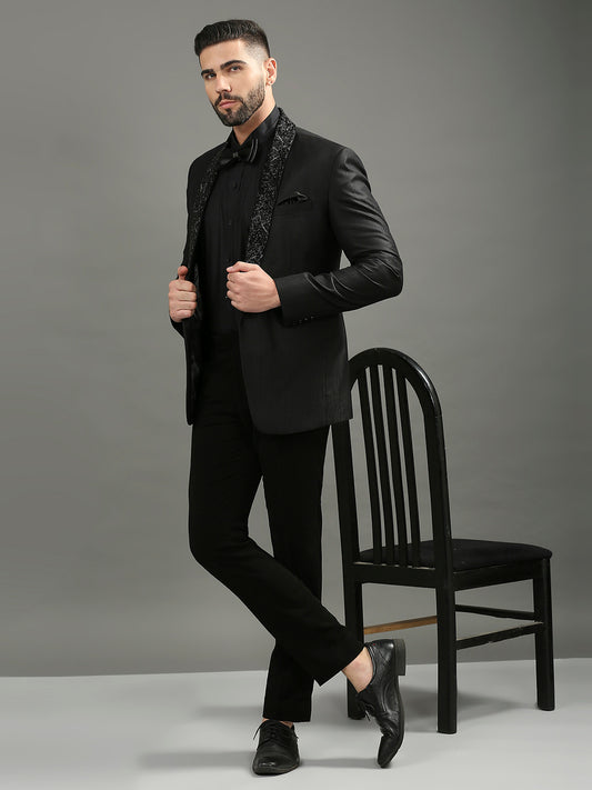 A man models a black men's formal suit consisting of a satin jacket with black patterned lapels, a black shirt, and black trousers, standing next to a black wooden chair against a gray studio background.