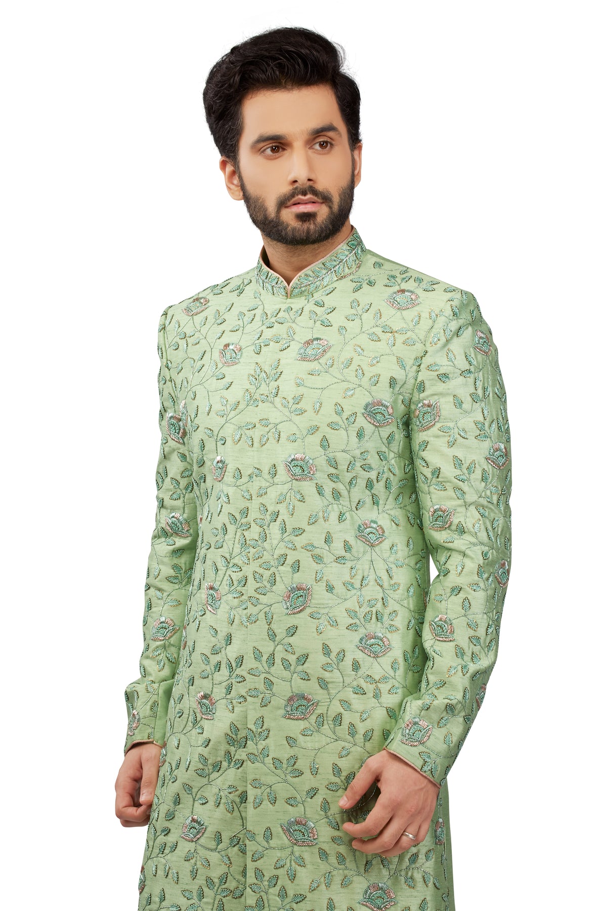 A man wearing a light green long-sleeve sherwani with an all-over floral and vine pattern in green, blue, and pink, photographed against a plain white background.