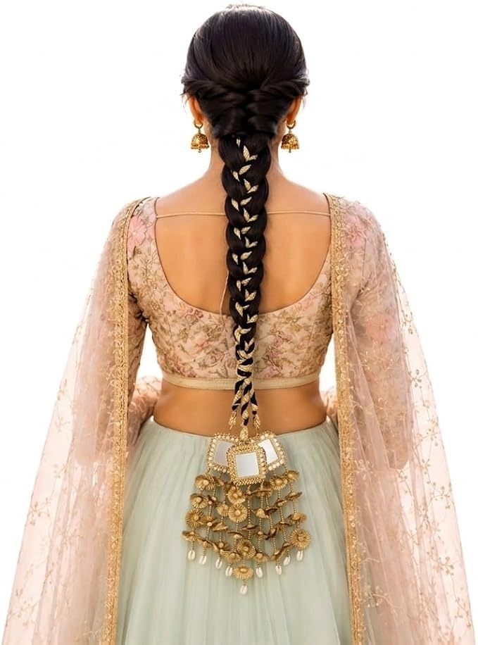Rear view of a woman wearing a traditional Indian outfit with a long dark braid adorned with gold coins, a light green skirt, and a beige floral embroidered blouse with sheer sleeves.