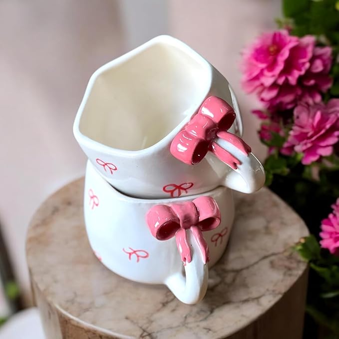 Two white ceramic cups with pink bow handles on a marble surface with flowers in the background.