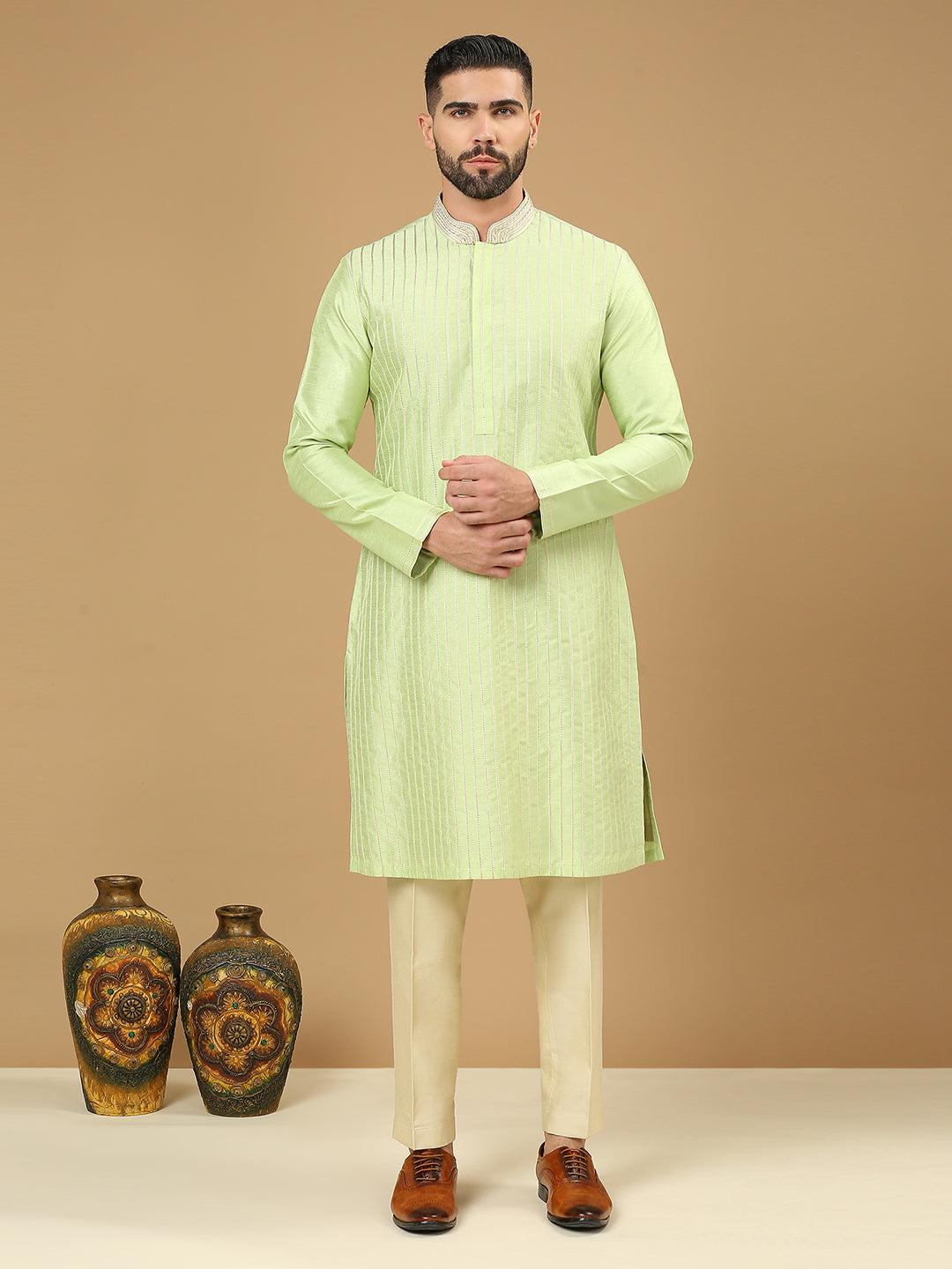 A man modeling a light green long-sleeve kurta with vertical texture, paired with cream trousers and brown shoes, standing against a solid beige studio background with two decorative vases on the floor.