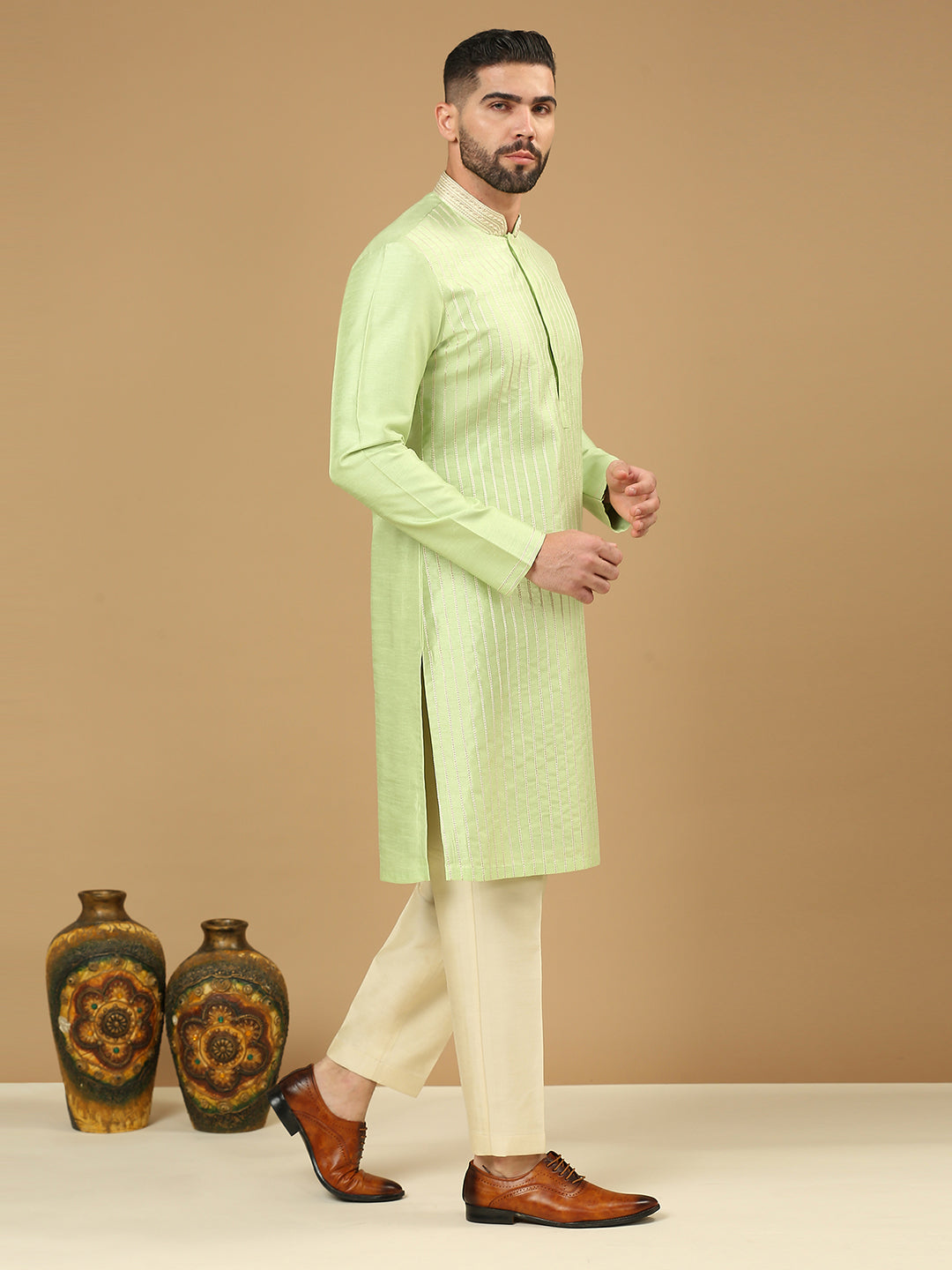 A man modeling a light green long-sleeve kurta with vertical stripes, paired with cream trousers and brown leather shoes, standing against a solid beige background next to two decorative vases.