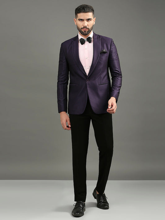 A man modeling a purple single-breasted tuxedo jacket with black trousers, a light pink dress shirt, a black bow tie, and a black pocket square, standing against a solid gray studio background.
