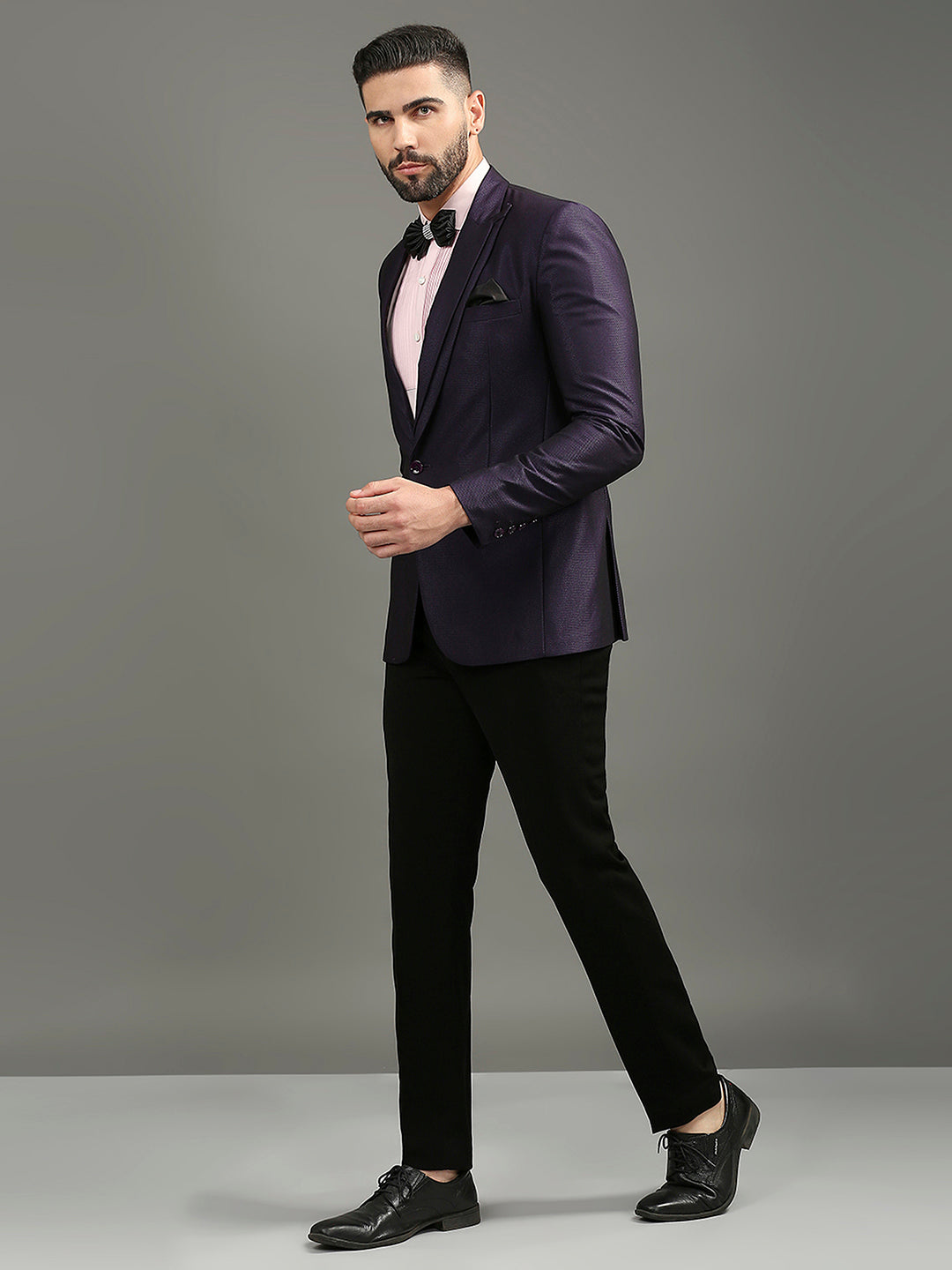 A man modeling a dark purple slim-fit tuxedo jacket with black satin peak lapels, paired with black trousers, a light pink dress shirt, a black bow tie, and black dress shoes, against a solid gray studio background.
