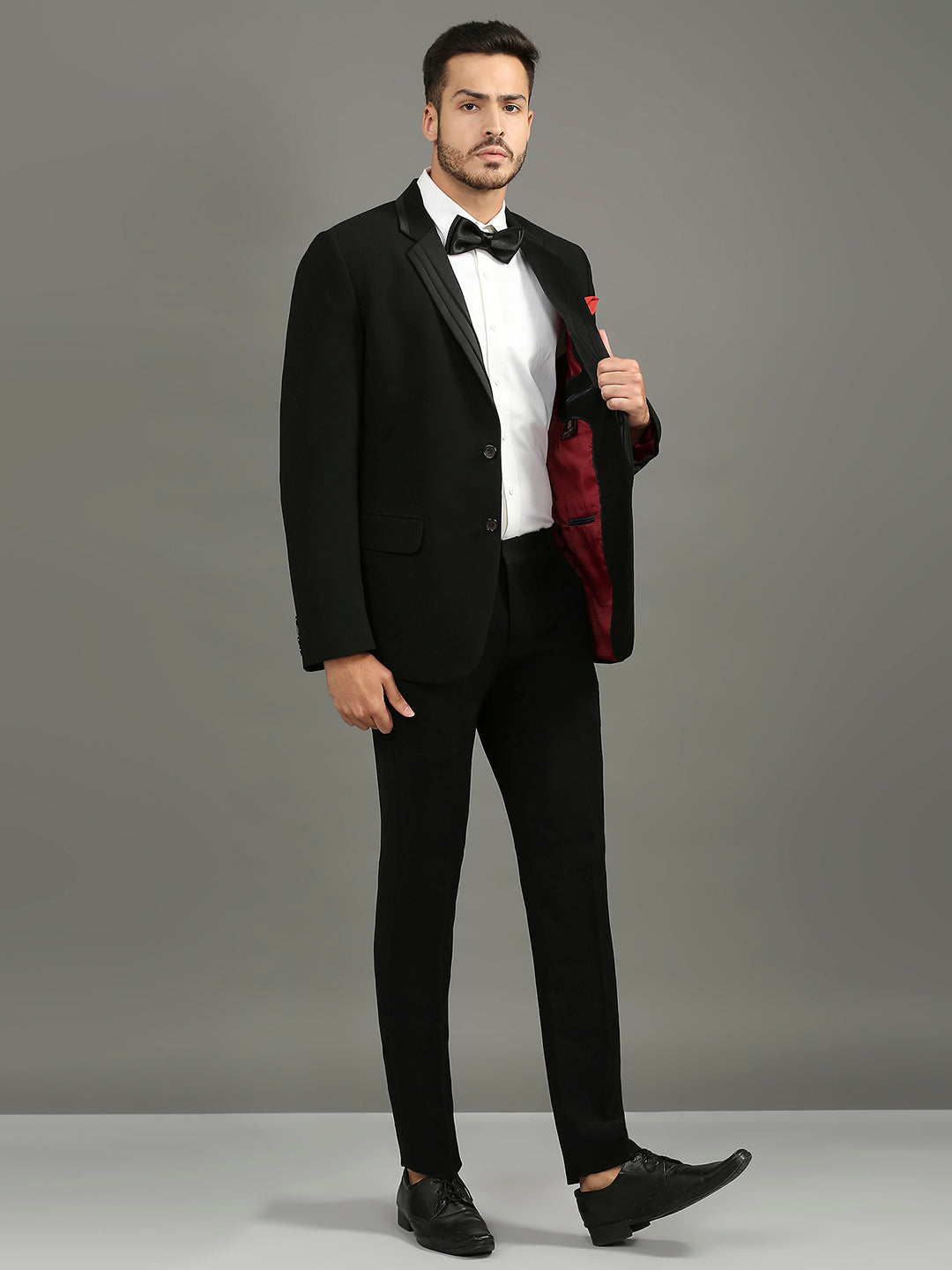 A man modeling a black tuxedo jacket with a white dress shirt, black bow tie, black trousers, and black dress shoes against a solid gray studio background.