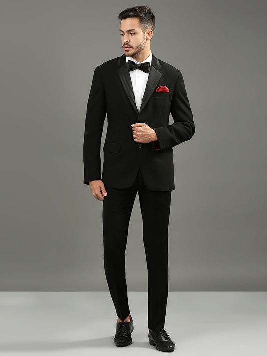 A man modeling a black tuxedo with black satin lapels, a white dress shirt, a black bow tie, and a red pocket square, standing against a solid gray studio background.
