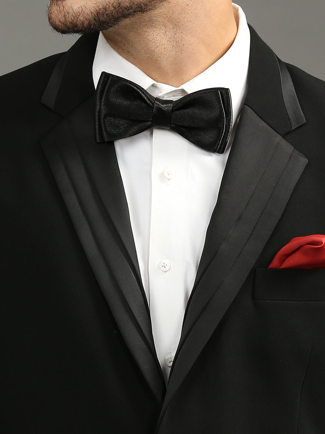 A close-up of a man wearing a black tuxedo jacket with satin lapels, a white dress shirt, a black velvet bow tie, and a red pocket square.