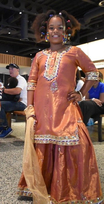 A woman modeling a traditional South Asian outfit in a metallic copper-orange color with intricate silver and gold embroidery, standing in an indoor event space.